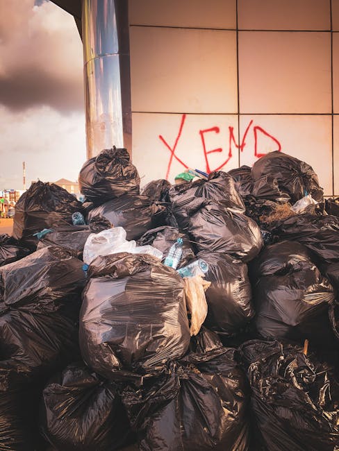 A large accumulation of black rubbish bags stacked against a modern building with a reflective, beige-colored tiled wall. The bags are filled with waste materials, with some transparent plastic bottles and other debris visible protruding from the openings. The surface of the bags appears crinkled and tied at the top, suggesting they are sealed for disposal. Behind the bags, there is red graffiti on the wall spelling out 'XEND.' The scene is outside, with part of the street or sidewalk visible at the base of the trash pile, and the lighting indicates late afternoon or early evening with overcast sky clouds. This image exemplifies an example of unmanaged waste, possibly highlighting the need for private rubbish collection or waste management services to prevent such urban clutter, supporting the context of alternative waste handling options often associated with independent rubbish removal in Elephant and Castle.