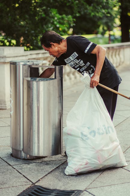 A middle-aged woman with short dark hair, wearing a black T-shirt with white text and dark trousers, is seen discarding a large white plastic bag filled with rubbish into a shiny stainless steel public litter bin on a paved sidewalk. The trash is situated in an outdoor area with a low stone balustrade and lush green trees in the background. The woman's right arm is extended as she holds the bag steady, and she appears focused on securely depositing the waste. The setting is daytime with natural light illuminating the scene, and the environment suggests a city or park frontage where independent waste disposal services might be utilized. The stainless steel litter bin has a rounded top with a narrow opening for depositing waste, consistent with urban rubbish collection standards. To the left of the woman, part of a wooden broom handle is visible, implying she may have been sweeping before disposal. The overall scene reflects a routine act of waste management by individuals, which ties into the context of private rubbish removal and alternative waste handling services that companies like wasteremovalelephantandcastle.co.uk provide in urban areas.