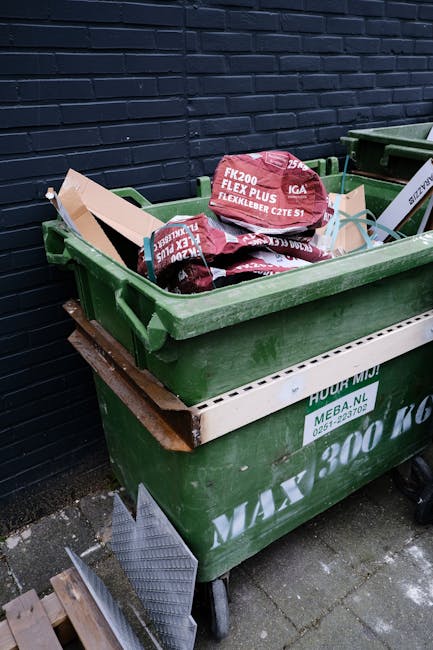 A large green wheelie bin filled with broken red and black plastic bags, positioned on a paved surface against a dark brick wall. The bags contain unspecified waste and are partially torn, with some packaging material visible. The bin has a white strip along the side with printed green text, indicating company details, and is situated next to another similar bin. The scene suggests an outside rubbish collection area, with the bins likely used for private waste disposal from nearby premises, reflecting alternative waste handling methods apart from local authority collection.
