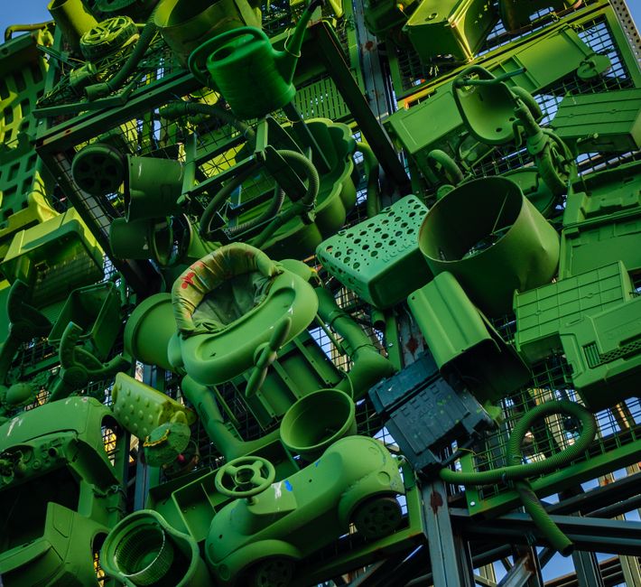 A large vertical structure composed of numerous green plastic objects, including chairs, buckets, pipes, and various household items, all densely stacked and attached to a metal framework. The objects display different textures, from smooth plastic surfaces to perforated and ribbed finishes, with some showing signs of wear and scratches. The grouping includes a children's high chair, garden pots, tool handles, and storage containers, arranged without apparent order. The environment appears to be indoors, possibly a warehouse or a dedicated waste storage area, illuminated by natural or artificial light that highlights the varied shades of green and shadows within the dense assembly. The scene closely relates to private waste handling or alternative rubbish collection methods, such as on-site clearance or waste collection for recycling, typical of services provided by companies specializing in rubbish removal and disposal solutions in urban settings.