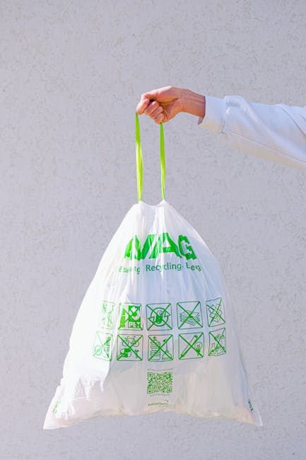 A human hand, partially visible and dressed in a white long-sleeve garment, is holding a white recycling bag by its bright green handles. The bag, made from thin, semi-transparent plastic, features green printed recycling symbols, text indicating its eco-friendly purpose, and various cleaning and recycling instructions arranged in a grid. The background consists of a plain, light grey wall with a smooth, slightly textured surface. The scene emphasizes the process of eco-conscious waste disposal or recycling, aligning with private waste management practices and promoting environmentally responsible rubbish handling. The overall lighting is neutral and evenly distributed, highlighting the details of the bag's material and printed symbols, fostering an image suitable for waste removal services focused on sustainable disposal methods.