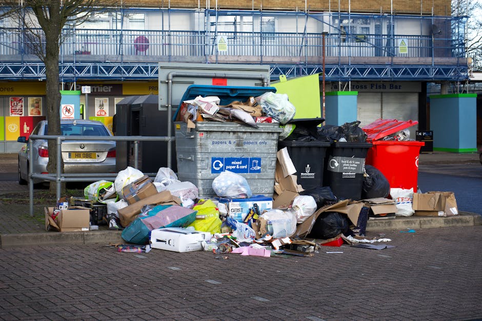A large collection of mixed household waste and clutter is piled around two open-top wheelie bins on a paved urban street. The waste includes crumpled cardboard boxes, plastic bags filled with垃圾, paper, and packaging materials, some spilling onto the surrounding pavement. The bins are positioned in front of a metal railing, with a compact silver car parked to the left. In the background, a building with blue scaffolding and a storefront with signage is visible, indicating ongoing work or renovation. The scene illustrates the type of informal rubbish accumulation that professional waste removal services, such as those provided by wasteremovalelephantandcastle.co.uk, frequently handle through private or alternative disposal methods, especially in areas where on-site clearance or independent collection is required.