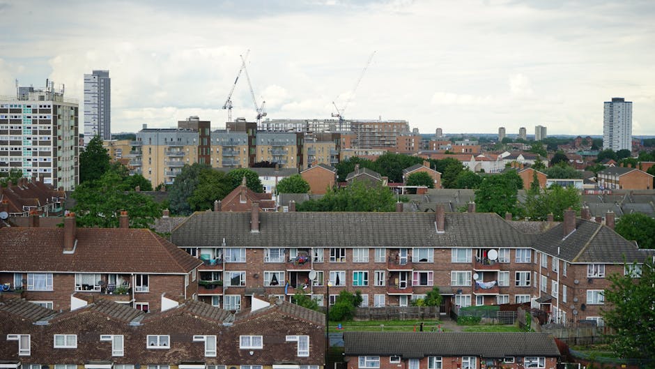 A panoramic view of an urban residential area showing multiple three-story brick apartment buildings with pitched tiled roofs in the foreground. Each building features small balconies with metal railings, some adorned with potted plants or chairs, and windows of various sizes, including bay and sliding styles. The buildings are separated by narrow pathways and small yards with green lawns and trees, creating a suburban feel within a cityscape. Beyond the residential area, there are several mid-rise and high-rise apartment blocks, some under construction with visible cranes and scaffolding, indicating ongoing development. The background skyline includes taller buildings against a cloudy sky, giving an industrial and evolving urban atmosphere. The environment appears well-maintained, with no visible litter, and the scene highlights a typical London or UK cityscape, which could be part of a property clearance or renovation project that might involve services like those offered by wasteremovalelephantandcastle.co.uk for private rubbish removal and on-site waste disposal.