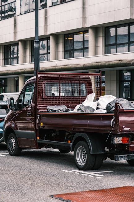 A small, dark red flatbed pickup truck is parked on an urban street in front of a modern multi-storey building with large glass windows and concrete panels. The truck's cargo area is filled with various types of rubbish, including large black and white refuse bags, some appearing partially open with contents inside, and other debris. The vehicle features a metal protective grid at the cab's rear, with the rubbish piled behind it, extending to the tailgate. Surrounding the truck, there are other parked vehicles visible, and a black lamppost stands nearby, partially obscuring part of the building. The scene appears to be during daylight with natural lighting, and the overall environment suggests a city setting where private waste collection services, such as those offered by waste removal companies, are operating to handle domestic and commercial rubbish, supporting alternative waste management solutions outside of local authority collection.