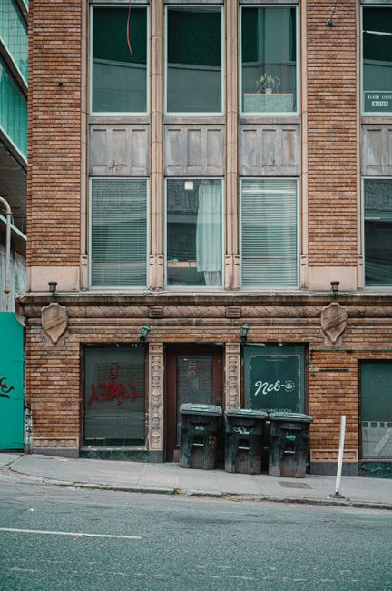 A multi-story brick building with large front windows featuring metal and glass frames, some with closed blinds or curtains. The ground floor has a central entrance door with a dark finish, flanked by two glass storefronts with graffiti on the left and right sides. In front of the building, on the sidewalk, there are three black wheelie bins and one smaller grey bin positioned against the wall, indicating private waste collection. To the right, a white pollard post is visible on the street edge. The pavement is concrete, and the street surface appears tarmac. The scene is illuminated naturally, with a neutral urban atmosphere suitable for material handling or rubbish removal services related to property clearance or on-site waste management, as sometimes performed independently of local authority collection by waste removal specialists like wasteremovalelephantandcastle.co.uk.