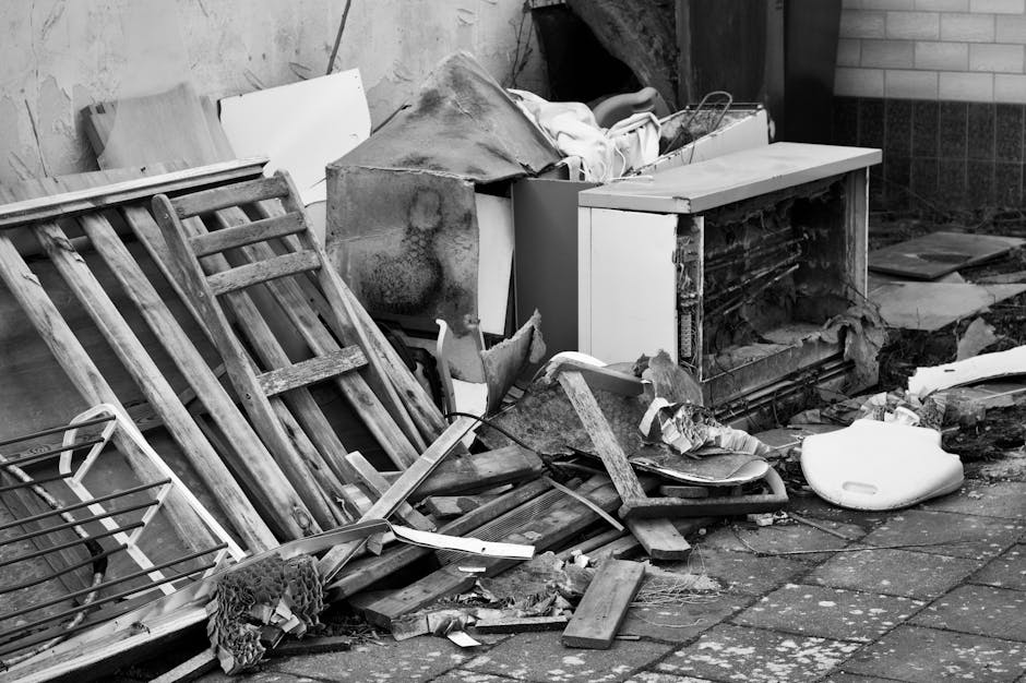 The image depicts a cluttered outdoor area near a building wall, featuring a disorganized pile of discarded items including a wooden slatted bed frame leaning against a brick wall, with visible weathering and peeling paint. Adjacent to it is a broken white kitchen drawer unit with its front panel missing, revealing scratched, exposed internal structures. On top of the unit, there are miscellaneous objects such as a tarp partially draped over the pile, a white cloth, and some small debris. Scattered across the ground are broken wooden planks, a twisted piece of metal, and a white plastic object that appears to be a displaced household item. A few tiles and debris are spread on the paved surface, which consists of dark stone slabs. The scene reflects typical private waste accumulation, possibly awaiting removal, indicative of a waste management or rubbish removal scenario handled by companies like Waste Removal Elephant and Castle. The lighting suggests an overcast day with diffused natural light illuminating the scene evenly, emphasizing the textures and condition of the discarded materials.